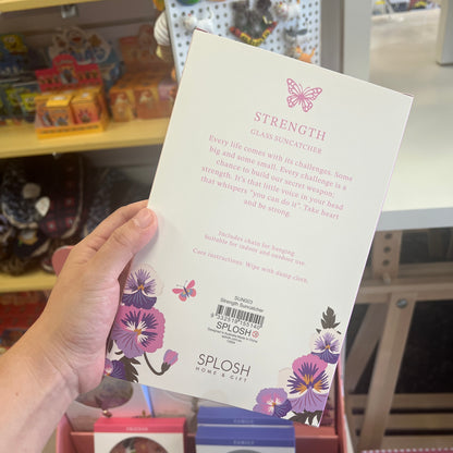 A hand holds a white card back for a SPLOSH Strength Glass Suncatcher, showing the product details, text instructions, and purple flower illustrations, with a blurred store background.