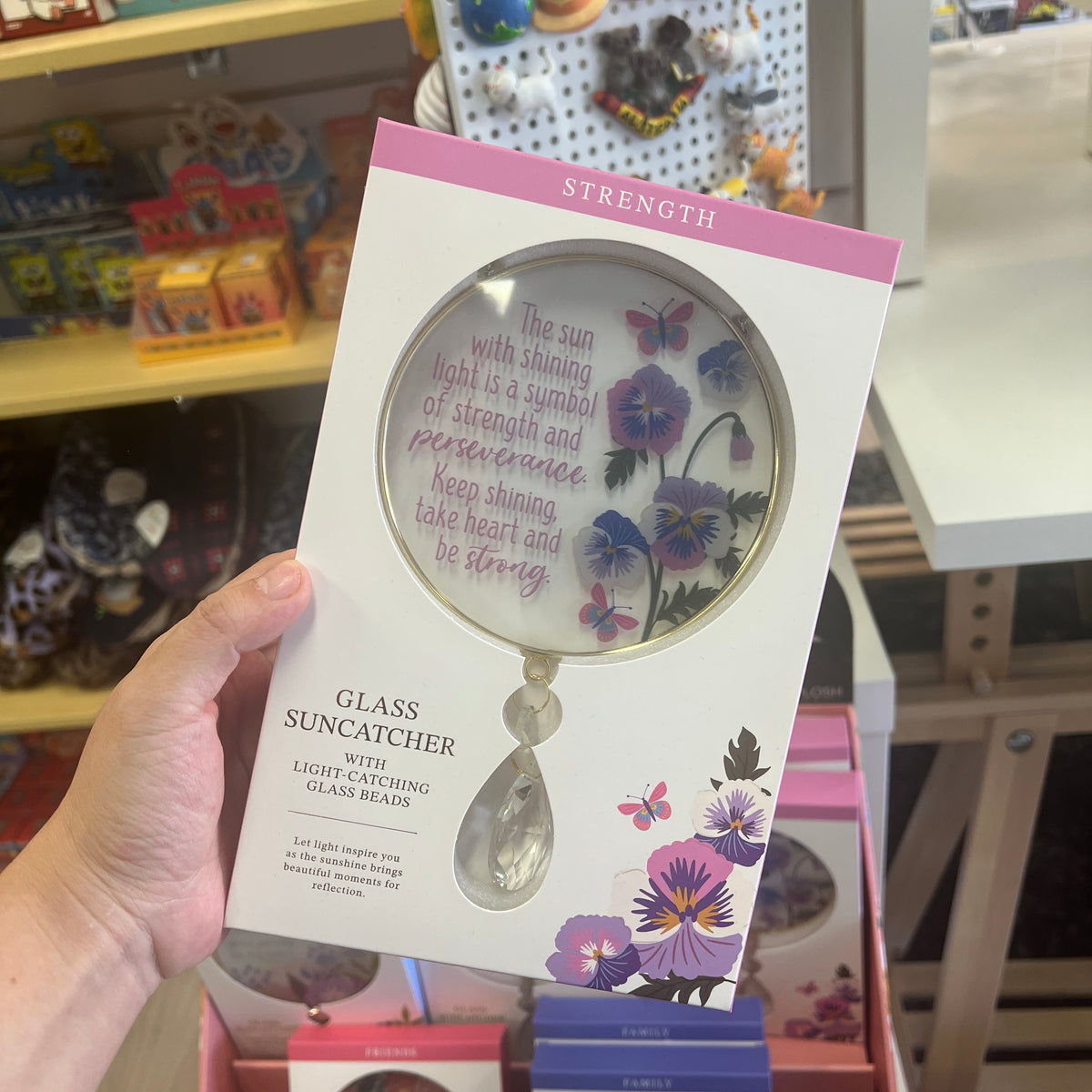 A hand holds a packaged glass suncatcher in a retail store. The product is a clear glass suncatcher with purple and pink floral illustrations and inspirational text on the circular mirror, presented in a white box with pink accents.