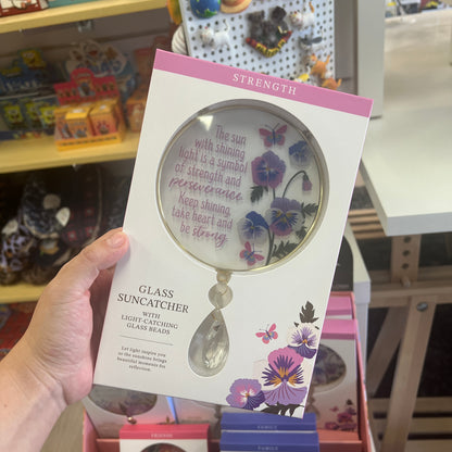 A hand holds a packaged glass suncatcher in a retail store. The product is a clear glass suncatcher with purple and pink floral illustrations and inspirational text on the circular mirror, presented in a white box with pink accents.