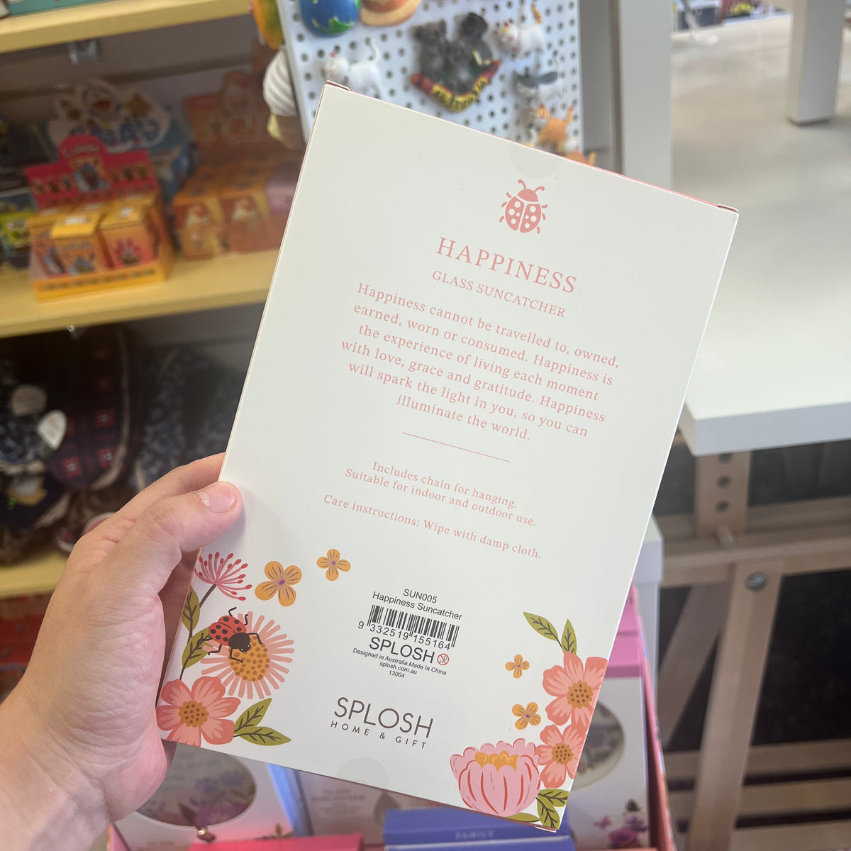 A hand holds a back view of the SPLOSH Home & Gift Happiness Glass Suncatcher packaging in a retail store setting. The packaging is cream-colored with pink and orange floral illustrations and text detailing the product features.