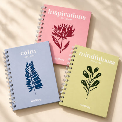 Three spiral-bound 2027 diaries from WellBeing arranged on a light beige surface with soft shadows. The diaries have pastel covers in light blue, pink, and pale yellow, each with a different botanical illustration and the words 'calm,' 'inspirations,' and 'mindfulness' on the front.