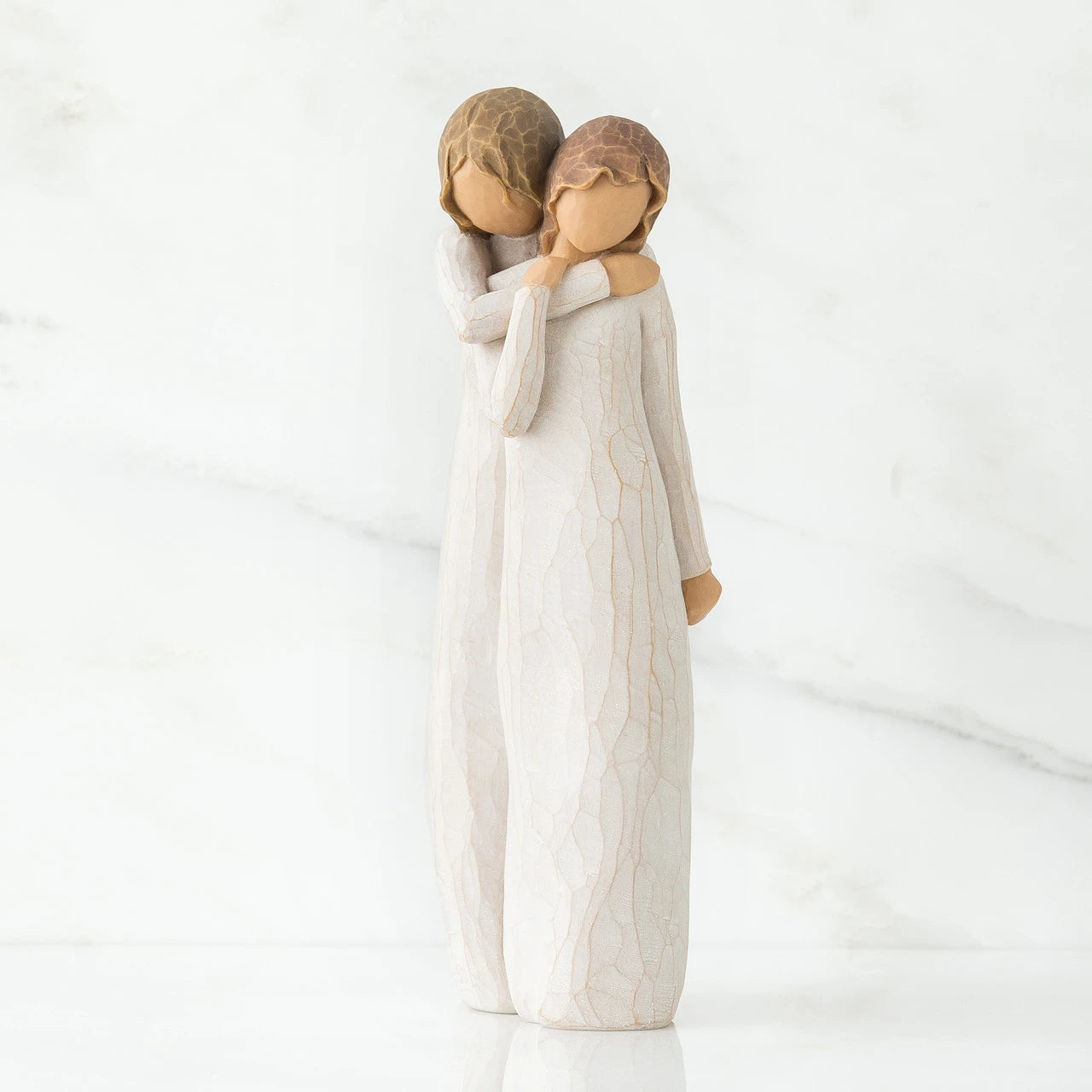 A Willow Tree figurine of a mother and daughter embracing, standing on a white reflective surface against a white marble background.