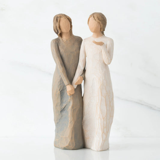 A pair of Willow Tree my sister my friend figurines depicting two women holding hands, one with a textured gray-brown dress and the other with a textured off-white dress, set against a clean white marble background.
