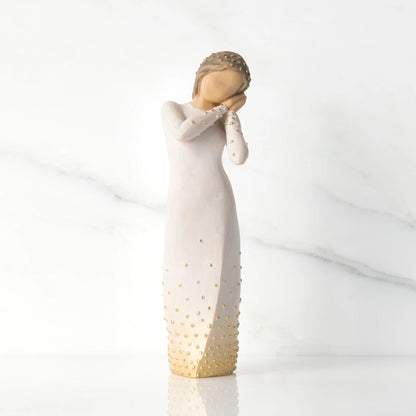 A Willow Tree wishing figurine of a woman in a long dress with gold studded details, standing on a reflective white surface against a white marble background.