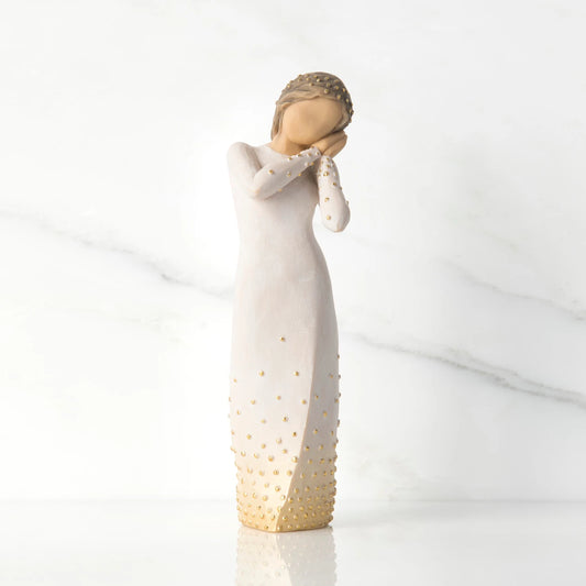 A Willow Tree wishing figurine of a woman in a long dress with gold studded details, standing on a reflective white surface against a white marble background.